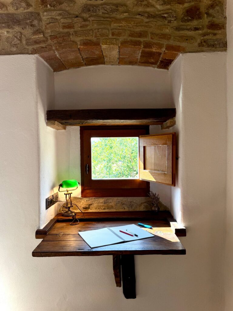 Scrittoio antico in legno ricavato in una nicchia sotto una finestra medievale, con lampada ministeriale verde e vista sulla campagna umbra: l'angolo perfetto per la lettura e lo smart working alla Torre della Soara.
