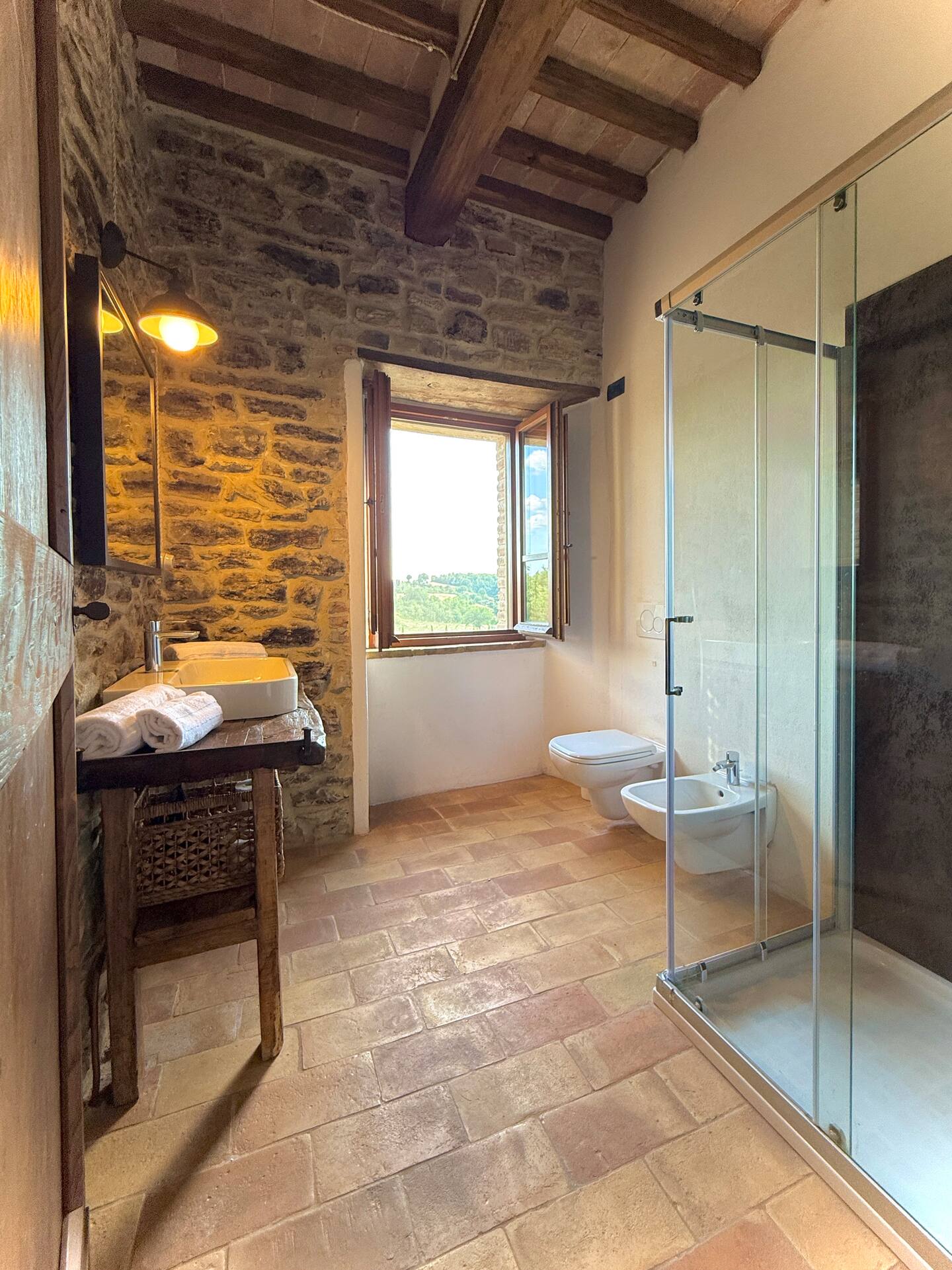 Vista panoramica del bagno alla Torre della Soara: cabina doccia moderna in vetro, sanitari sospesi bianchi e lavabo su mobile in legno antico, con una finestra aperta che incornicia il paesaggio collinare umbro. Vista panoramica del bagno alla Torre della Soara: cabina doccia moderna in vetro, sanitari sospesi bianchi e lavabo su mobile in legno antico, con una finestra aperta che incornicia il paesaggio collinare umbro.