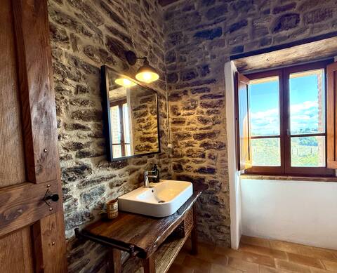 Dettaglio del bagno della Torre della Soara: lavabo moderno su mensola in legno antico, specchio con illuminazione vintage e finestra aperta con vista panoramica sulle verdi colline umbre. Dettaglio del bagno della Torre della Soara: lavabo moderno su mensola in legno antico, specchio con illuminazione vintage e finestra aperta con vista panoramica sulle verdi colline umbre.