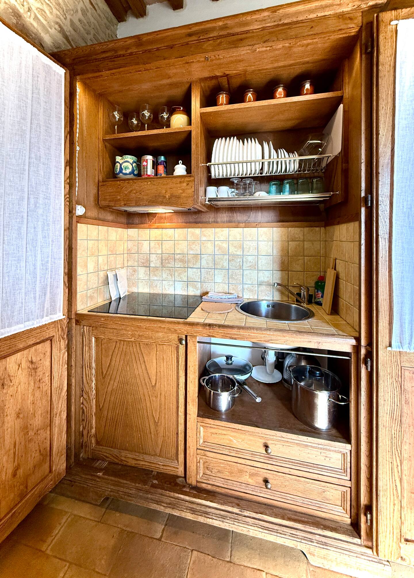 Dettaglio della cucina attrezzata a scomparsa della Torre della Soara: piano cottura a induzione, lavello in acciaio, stoviglie ordinate su mensole in legno e set di pentole, tutto integrato in un armadio rustico. Dettaglio della cucina attrezzata a scomparsa della Torre della Soara: piano cottura a induzione, lavello in acciaio, stoviglie ordinate su mensole in legno e set di pentole, tutto integrato in un armadio rustico.