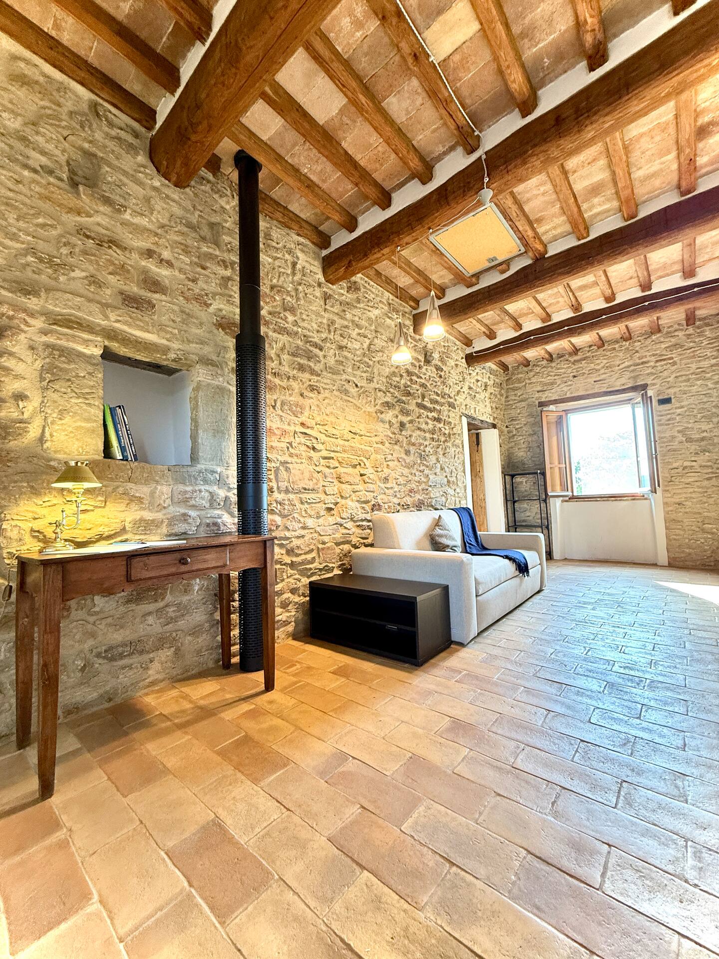 Zona living della Torre della Soara con pareti in pietra originale, soffitto a travi, nicchia libreria illuminata da una lampada classica su scrivania in legno e divano moderno. Zona living della Torre della Soara con pareti in pietra originale, soffitto a travi, nicchia libreria illuminata da una lampada classica su scrivania in legno e divano moderno.