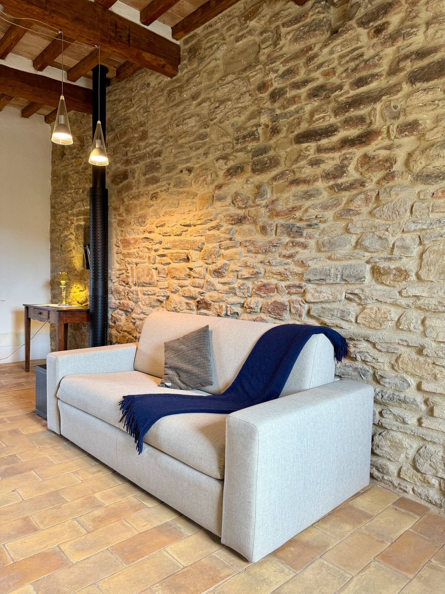 Dettaglio zona living alla Torre della Soara: divano moderno color sabbia con plaid blu, accostato a una parete in pietra antica illuminata da lampade a sospensione di design. Dettaglio zona living alla Torre della Soara: divano moderno color sabbia con plaid blu, accostato a una parete in pietra antica illuminata da lampade a sospensione di design.