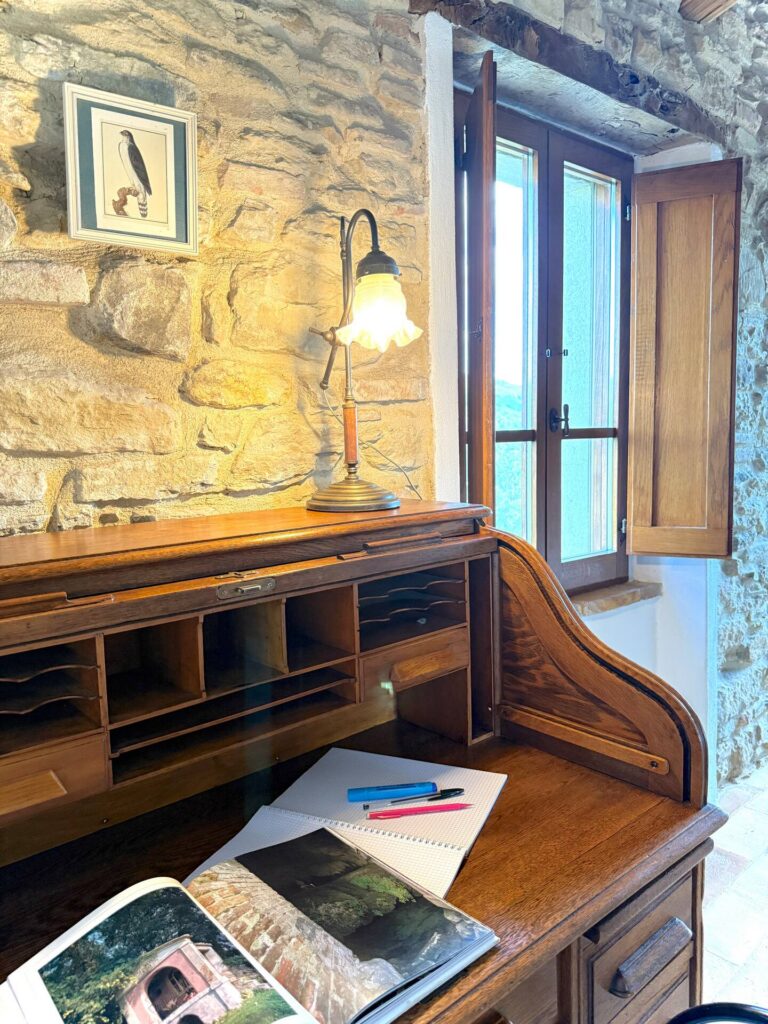Scrivania secretaire d'epoca in legno nobile nella camera dell'appartamento grande, accostata a una parete in pietra originale con lampada vintage accesa e finestra panoramica.