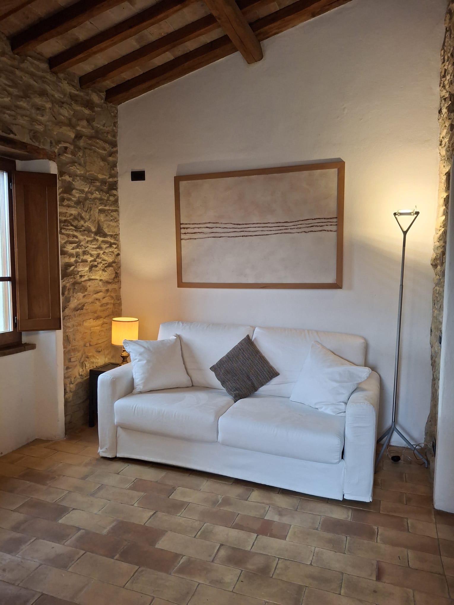Zona relax dell’appartamento Torre alla Torre della Soara, con divano chiaro, parete in pietra a vista, pavimento in cotto e soffitto con travi in legno.